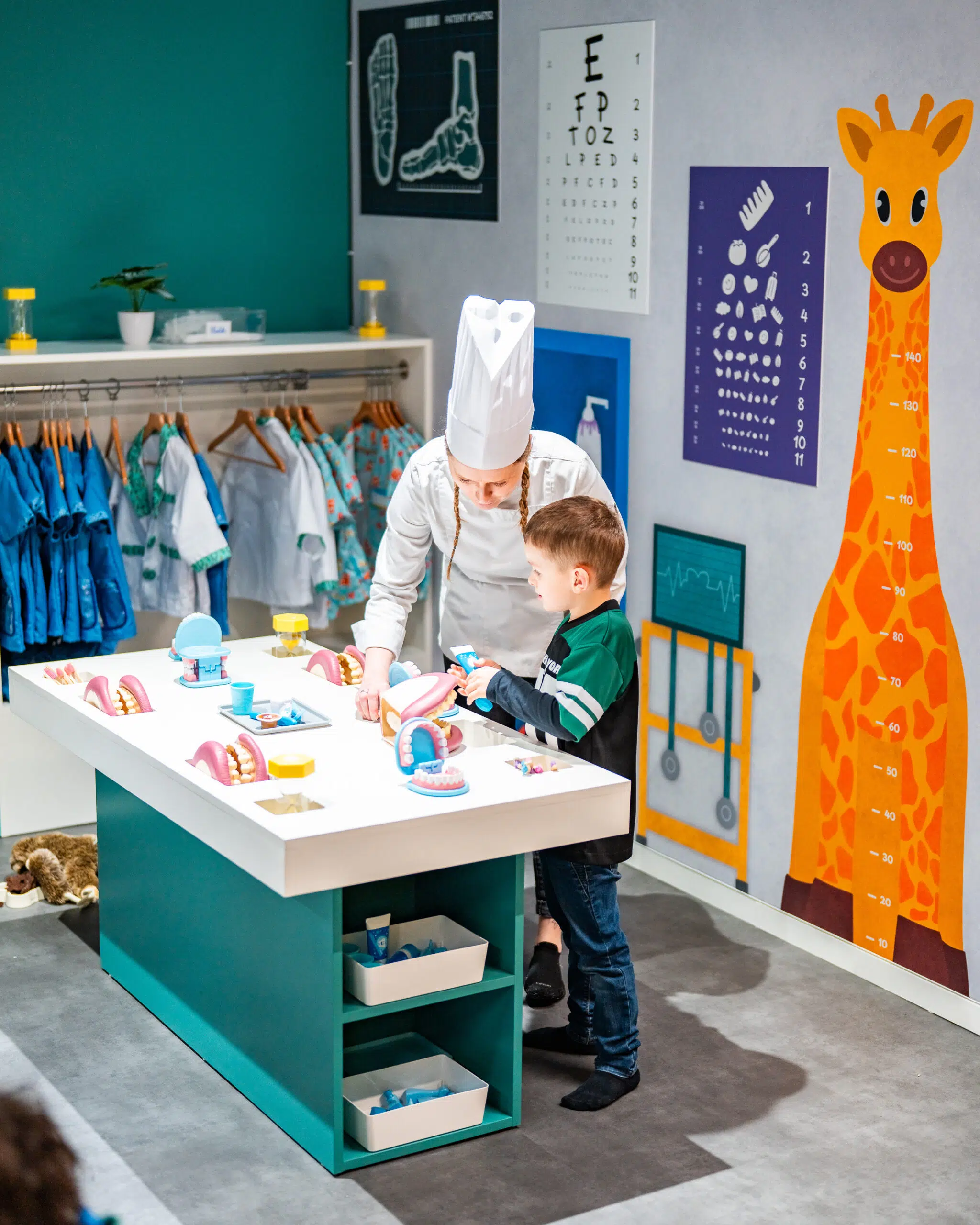 Enfants au frais dans le parc indoor climatisé Palomano Metz en été Un enfant participe à une activité de dentiste dans un espace immersif de Palomano Metz. Accompagné de sa maman déguisée en cuisiniere, il découvre le matériel dentaire sur une table de jeu. Le décor reproduit un véritable cabinet médical avec des blouses, des affiches pédagogiques, une toise en forme de girafe et différents accessoires adaptés aux enfants.