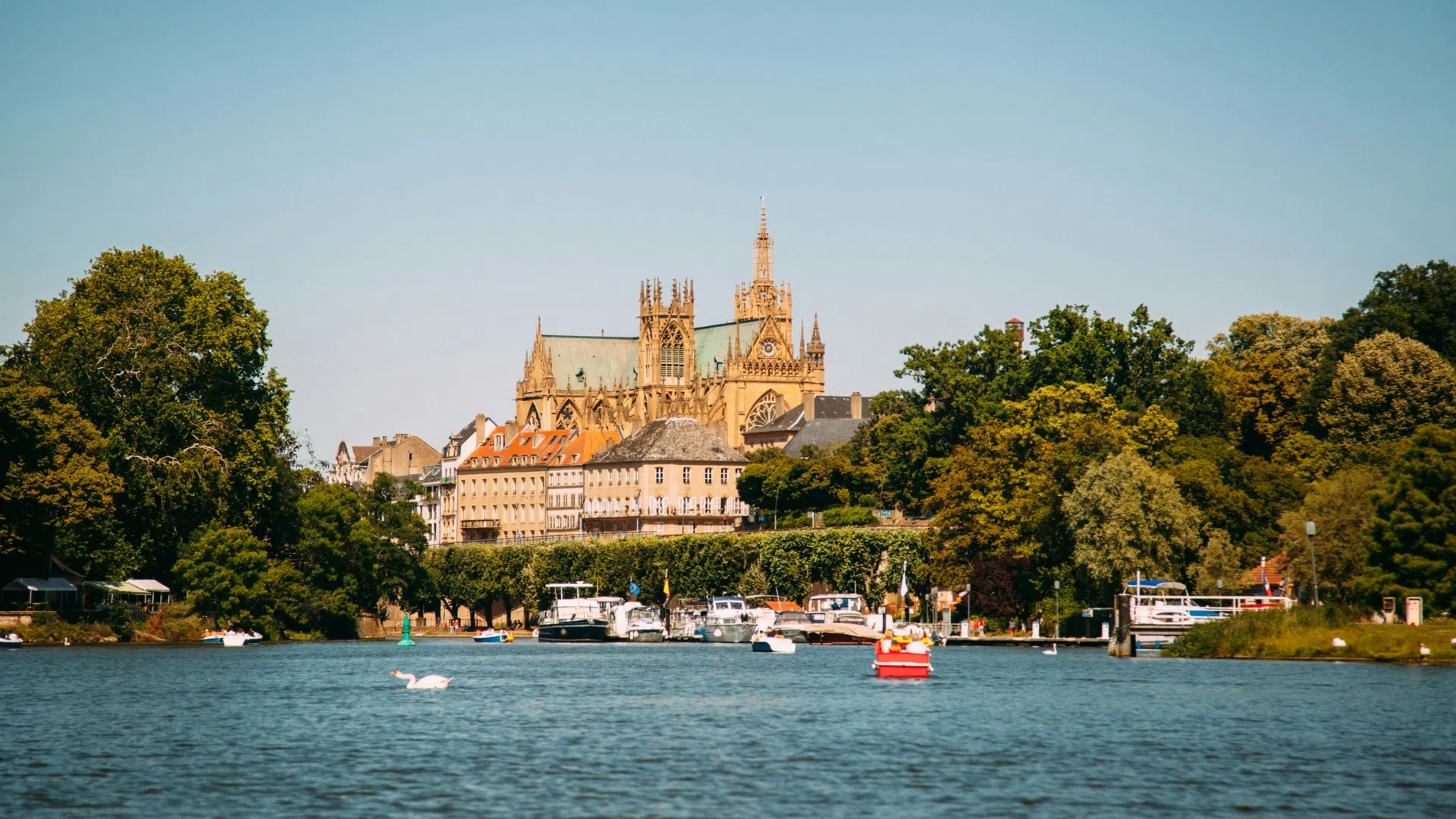 Sortie en famille avec enfants pendant les vacances d’été à Metz Vue panoramique sur le plan d'eau de Metz avec plusieurs bateaux amarrés et une embarcation navigant sur l'eau. En arrière-plan, la majestueuse cathédrale Saint-Étienne.